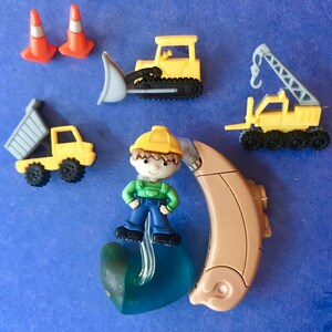 Pu&ograve; includere: Una figurina di un operaio edile blu e verde con un casco giallo, in piedi su un apparecchio acustico blu e verde. La figurina &egrave; circondata da veicoli da costruzione giocattolo, tra cui un bulldozer giallo, un camion ribaltabile giallo e una gru gialla.