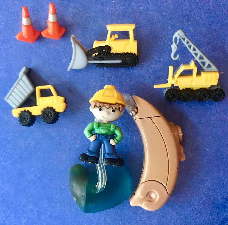 Pu&ograve; includere: Una figurina di un operaio edile blu e verde con un casco giallo, in piedi su un apparecchio acustico blu e verde. La figurina &egrave; circondata da veicoli da costruzione giocattolo, tra cui un bulldozer giallo, un camion ribaltabile giallo e una gru gialla.