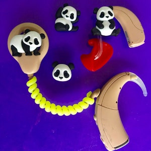 Puede incluir: Aud&iacute;fonos color beige con adornos de osos panda y un cable en espiral amarillo brillante. Los pandas son negros y blancos. Tambi&eacute;n hay un molde para el o&iacute;do rojo. El fondo es de color p&uacute;rpura liso.
