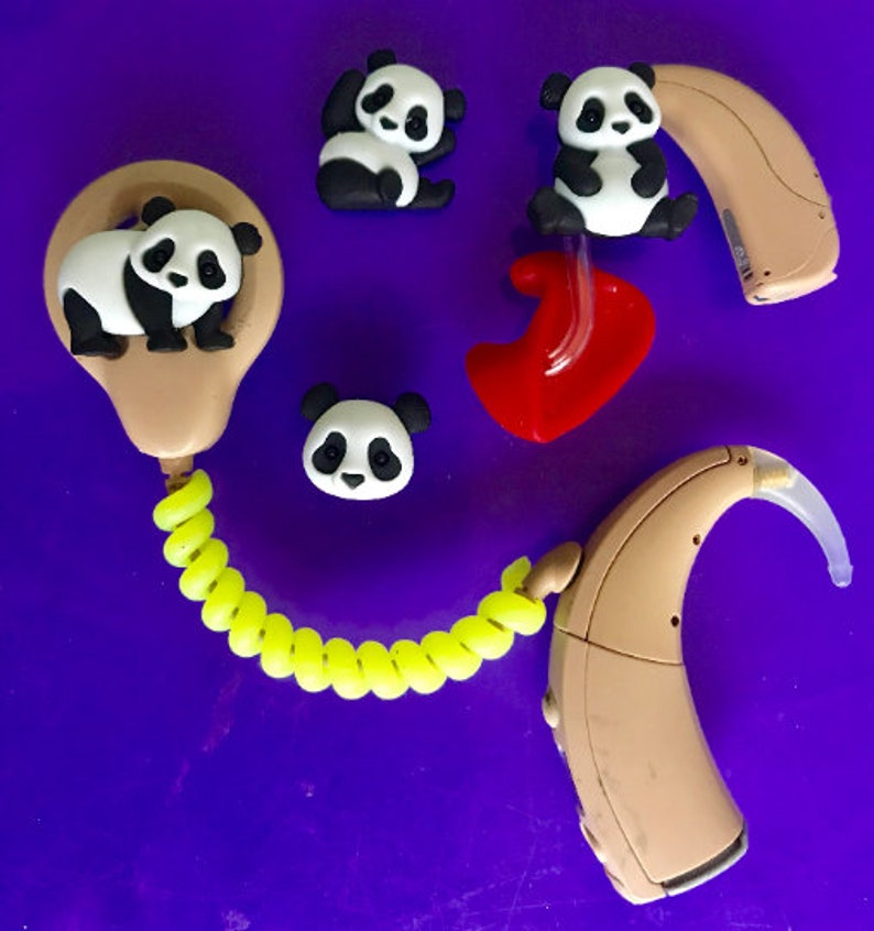 Puede incluir: Aud&iacute;fonos color beige con adornos de osos panda y un cable en espiral amarillo brillante. Los pandas son negros y blancos. Tambi&eacute;n hay un molde para el o&iacute;do rojo. El fondo es de color p&uacute;rpura liso.