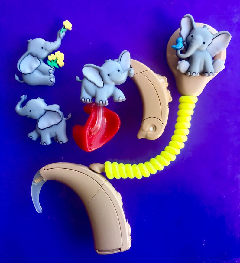Puede incluir: Una colecci&oacute;n de figuritas de elefantes grises con orejas grandes y un ribete azul claro, combinadas con aud&iacute;fonos beige. Un elefante sostiene flores, otro un coraz&oacute;n rojo y otro un p&aacute;jaro azul. Un tubo amarillo en espiral conecta los aud&iacute;fonos.