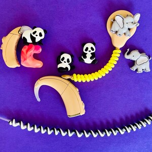 Puede incluir: Un conjunto de aud&iacute;fonos y accesorios, incluyendo amuletos con forma de panda y elefante, sobre un fondo morado. Los aud&iacute;fonos son beige, con un inserto rosa. Un cable amarillo en espiral se conecta a un molde para el o&iacute;do.