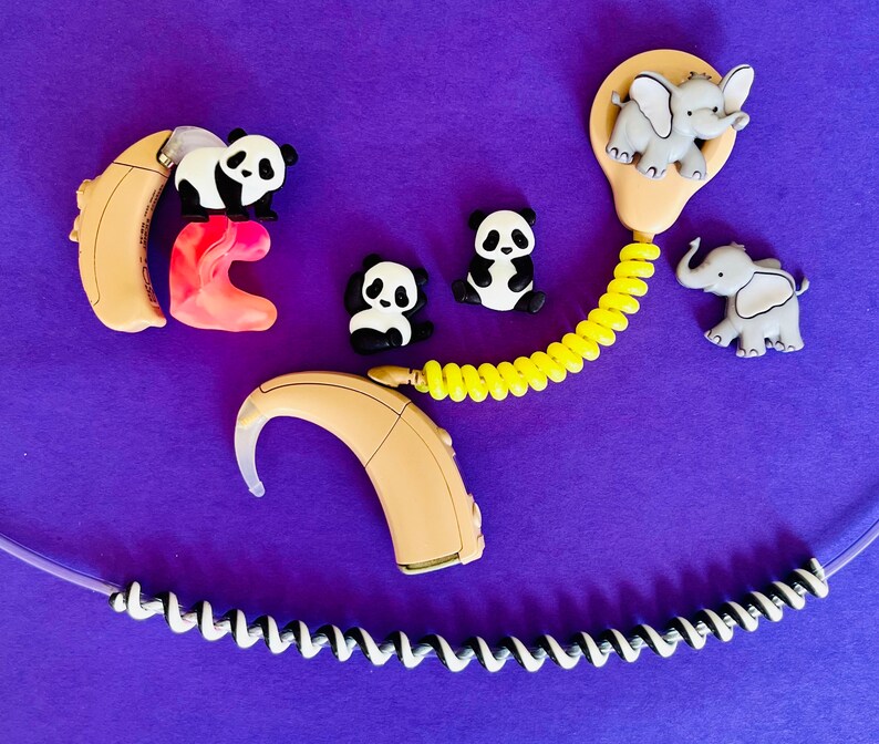 Puede incluir: Un conjunto de aud&iacute;fonos y accesorios, incluyendo amuletos con forma de panda y elefante, sobre un fondo morado. Los aud&iacute;fonos son beige, con un inserto rosa. Un cable amarillo en espiral se conecta a un molde para el o&iacute;do.