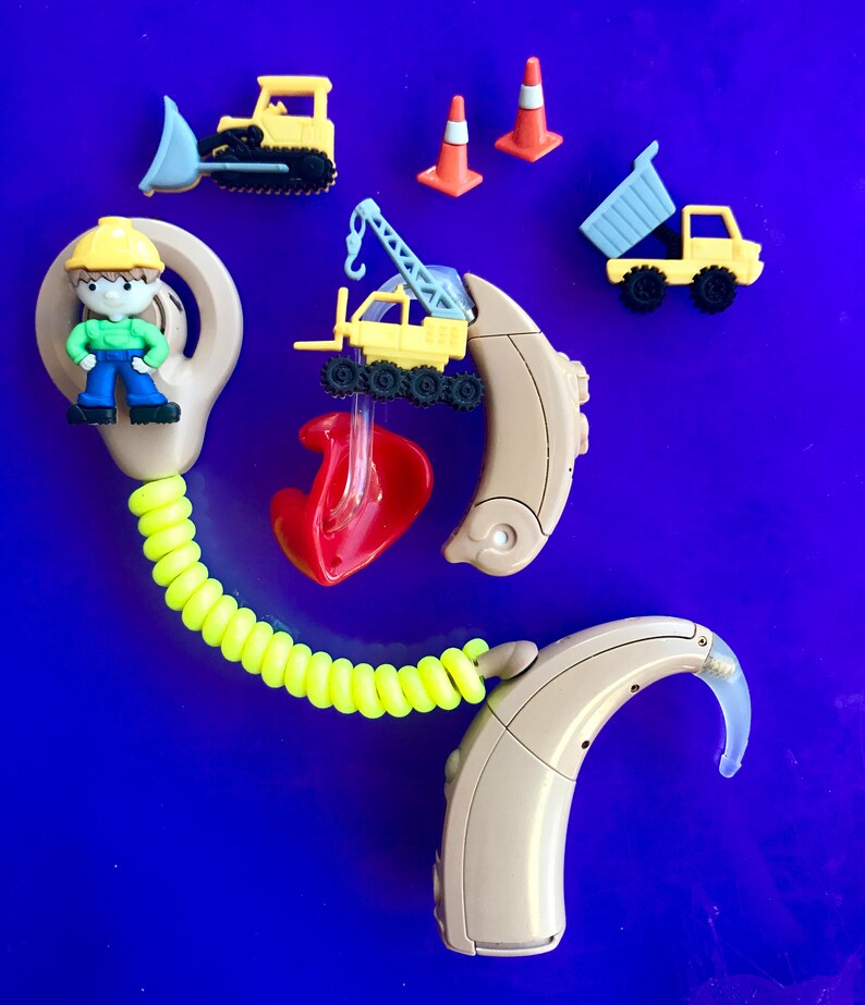 Pu&ograve; includere: Un apparecchio acustico beige con un cavo a spirale giallo e un'impronta auricolare rossa. L'apparecchio acustico &egrave; decorato con piccoli veicoli da costruzione giocattolo, tra cui un bulldozer giallo, una gru gialla, un camion ribaltabile grigio e due coni di segnalazione arancioni.