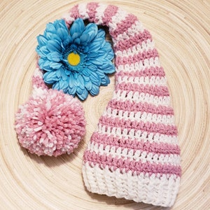 May include: A crochet hat featuring horizontal stripes of white and dusty pink yarn with a subtle metallic shimmer. The hat has a ribbed white brim, a long pointed shape, and a large, fluffy pom-pom attached to the side with a bright blue flower.