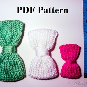 May include: Three knitted bow ties in different colors: green, white, and pink. The bow ties are arranged in a row from left to right, with the green bow tie on the left, the white bow tie in the middle, and the pink bow tie on the right. The bow ties are all made with a similar knit stitch pattern.
