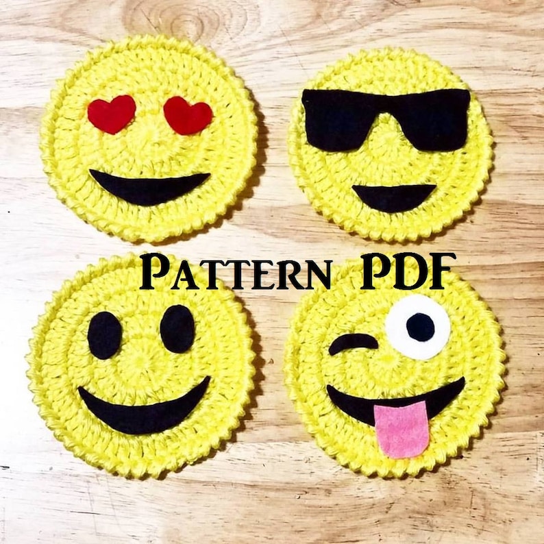 May include: Four crocheted yellow emoji coasters with different facial expressions. The coasters feature black felt eyes, mouths, and sunglasses. One coaster has red felt hearts for eyes. Another coaster has a white felt eye and a pink felt tongue. The coasters are on a wooden surface. PATTERN PDF is written in black text on the surface.