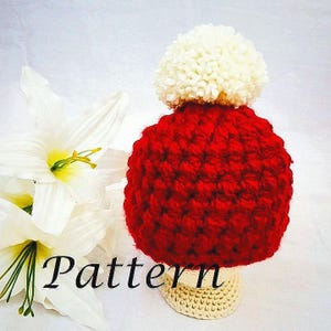 May include: A red crocheted hat with a white pom pom on top. The hat is sitting on a crocheted base.