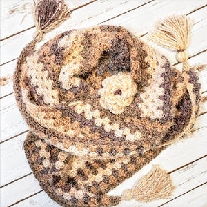 Puede incluir: Un chal de ganchillo hecho a mano en tonos marrón, beige y morado. El chal presenta un adorno floral y borlas decorativas en los extremos. El patrón de granny square crea un aspecto texturizado y acogedor.