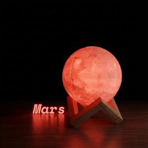 May include: A red Mars-themed lamp with a textured surface, resting on a wooden stand. The word "Mars" is illuminated in red. The lamp emits a warm, reddish glow against a dark background.