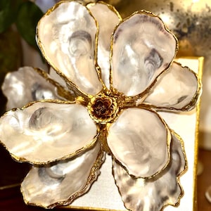 May include: A decorative flower made from oyster shells with gold-painted edges. The flower has a dark brown center and is mounted on a square, gold-edged base. The petals are white and pearlescent.