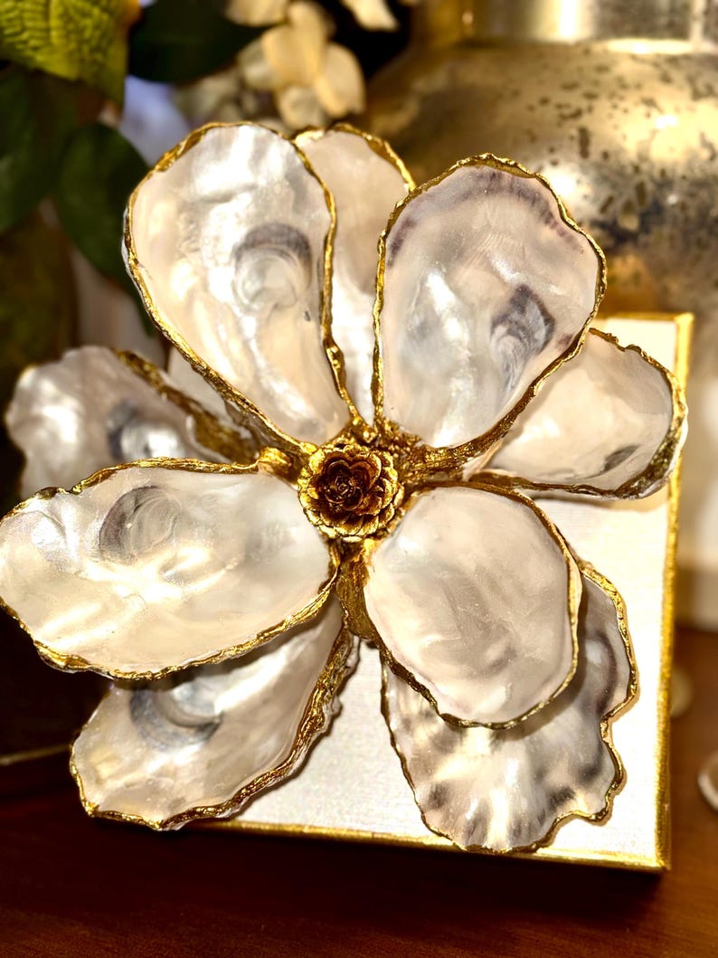 May include: A decorative flower made from oyster shells with gold-painted edges. The flower has a dark brown center and is mounted on a square, gold-edged base. The petals are white and pearlescent.