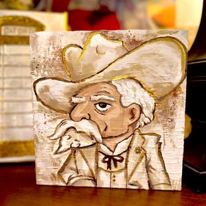 May include: A hand-painted artwork on a wooden block depicts a cartoonish cowboy with a white mustache and hat. The cowboy wears a white suit with gold accents. The background is a textured, off-white color.