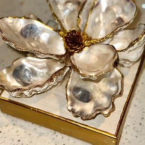 May include: A decorative flower made from oyster shells with gold trim, centered with a small pinecone. The flower sits atop a square, off-white surface with a gold border. The oyster shells have a pearlescent sheen.