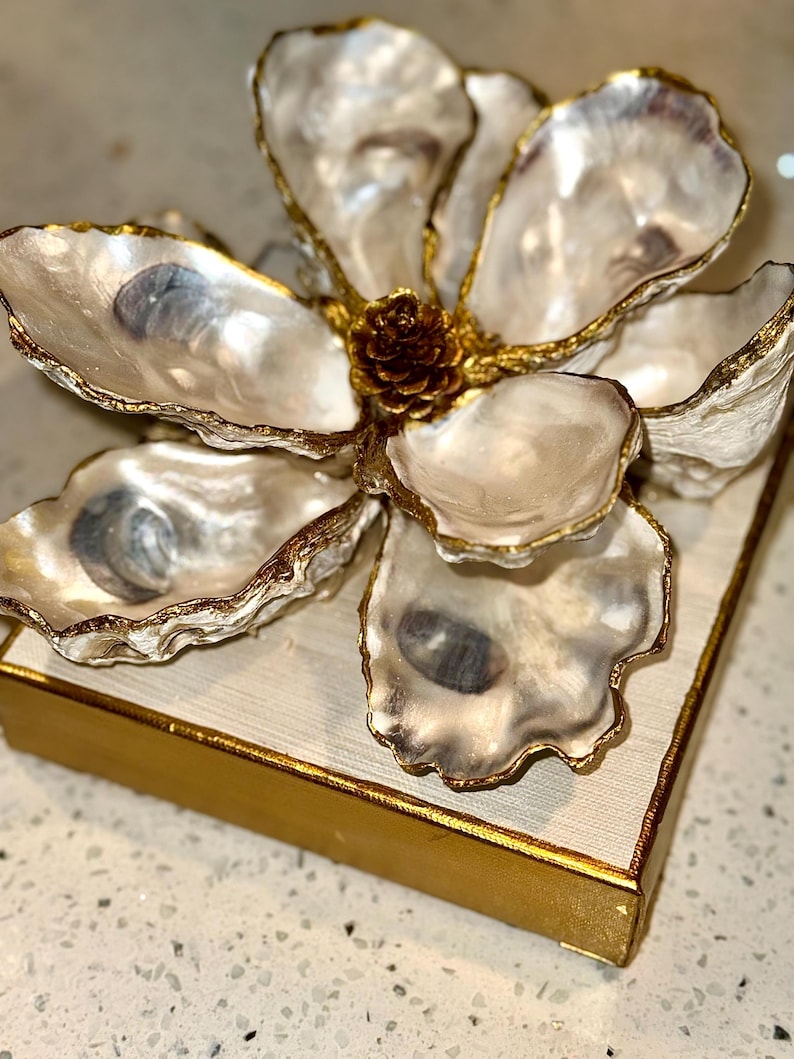 May include: A decorative flower made from oyster shells with gold trim, centered with a small pinecone. The flower sits atop a square, off-white surface with a gold border. The oyster shells have a pearlescent sheen.