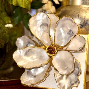 May include: A decorative flower made from white oyster shells with gold trim. The flower is mounted on a square, off-white canvas with gold edges. The center of the flower is a small, brown, textured element.