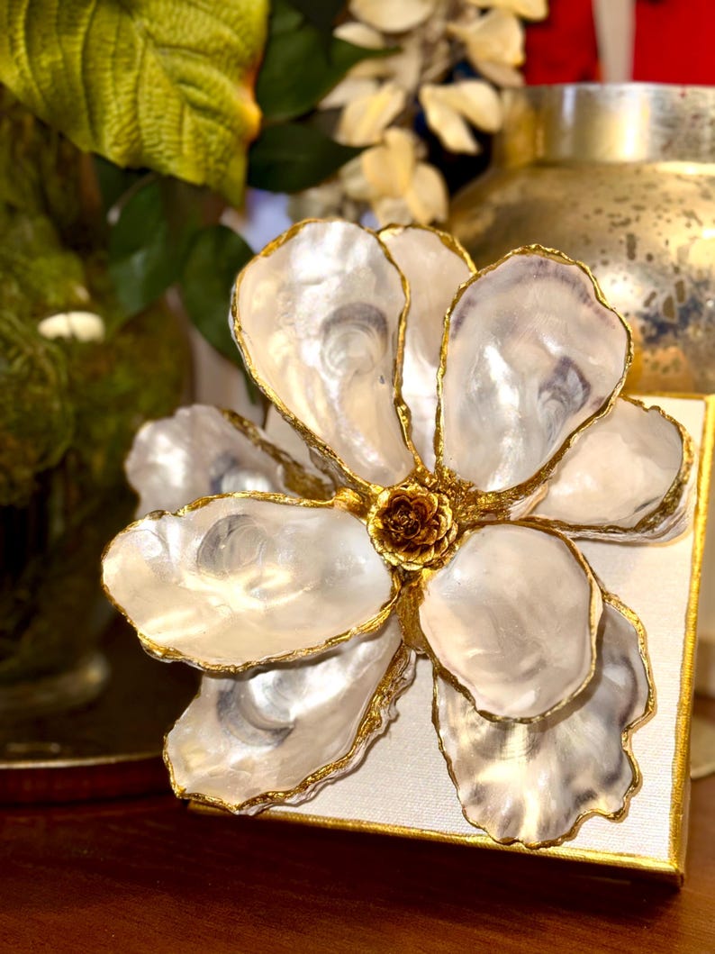 May include: A decorative flower made from white oyster shells with gold trim. The flower is mounted on a square, off-white canvas with gold edges. The center of the flower is a small, brown, textured element.