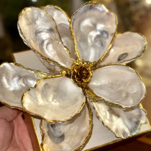May include: A decorative flower sculpture crafted from oyster shells. The petals are pearlescent white with gold-painted edges, arranged around a central, textured, golden core. The flower is mounted on a white square base with gold trim.