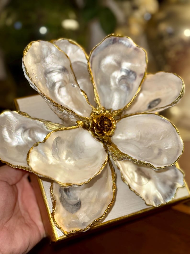 May include: A decorative flower sculpture crafted from oyster shells. The petals are pearlescent white with gold-painted edges, arranged around a central, textured, golden core. The flower is mounted on a white square base with gold trim.
