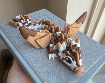 May include: A whimsical, handcrafted dragon figurine made from polymer clay. The dragon features a white and brown head, wings, and body, with details resembling graham crackers, chocolate, and marshmallows. It rests on a blue book with a decorative pattern.