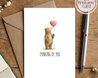 Pode incluir: Um cartão de felicitações branco com a ilustração de um urso castanho a segurar um balão vermelho em forma de coração com o texto "THINKING OF YOU" impresso por baixo. O cartão é indicado como imprimível 5x7 numa superfície de madeira.