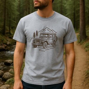 May include: A grey t-shirt with a brown line drawing of a vintage off-road vehicle, mountains, and trees. The short-sleeved shirt is made of a soft material, suitable for casual wear.