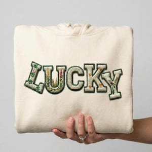 Peut inclure: Un sweat à capuche crème avec le mot "LUCKY" brodé sur le devant. Les lettres sont bordées de vert et remplies de motifs floraux. Le sweat à capuche est tenu dans une main.
