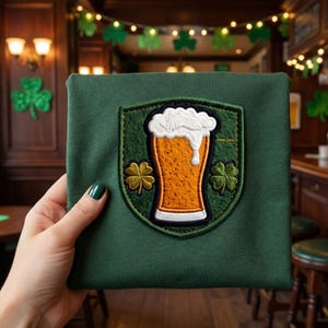Peut inclure: Un tissu carré vert avec un écusson brodé représentant un verre de bière avec de la mousse et des trèfles. Le tissu est tenu par une main avec du vernis à ongles vert. L'arrière-plan montre un cadre de bar avec des décorations de trèfles.
