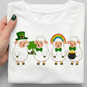 Peut inclure: T-shirt blanc avec un motif de la Saint-Patrick. Le motif présente quatre moutons de dessin animé, chacun avec un accessoire différent : un chapeau vert, un trèfle, un arc-en-ciel et une marmite d'or. Les moutons portent tous des nœuds papillon verts.