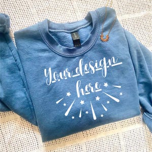 May include: A blue crewneck sweatshirt with the text "Your design here" in white script. The design includes star and burst graphics. A gold-colored necklace with a sun pendant is draped on the sweatshirt.