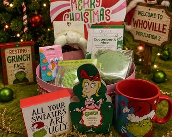Może przedstawiać: Świąteczny kosz prezentowy z czerwonym kubkiem z motywem Grincha, pudełkiem z napisem "All Your Sweaters Are Ugly" i znakiem "Welcome to Whoville". Kosz zawiera różne produkty kosmetyczne i dużą dekorację w paski z cukrowej laski.