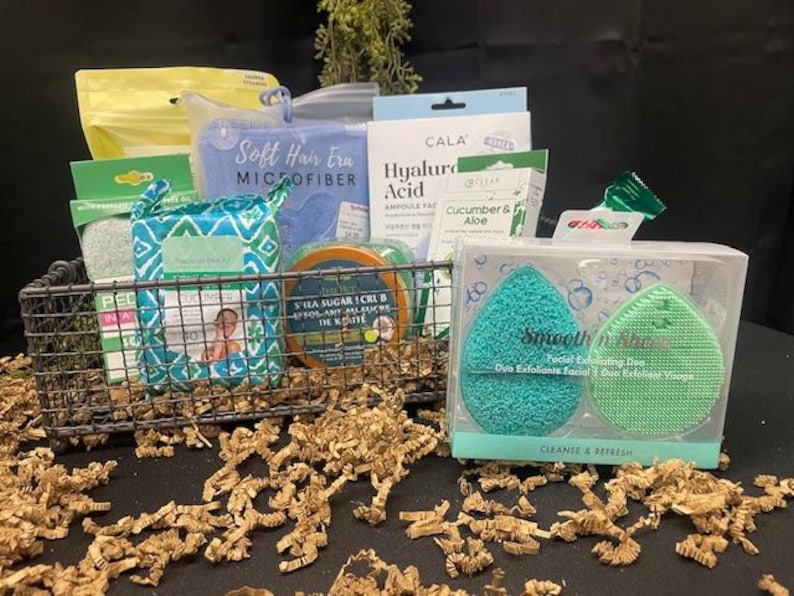 May include: A wire basket filled with skincare products, including a blue microfiber cloth, hyaluronic acid ampoules, and a duo exfoliating sponge set. The set includes a teal and green sponge. The basket is surrounded by brown paper.