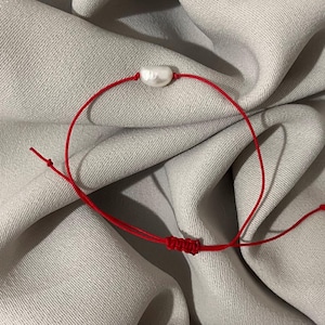 May include: A red string bracelet featuring a single, white, irregularly shaped pearl. The bracelet is knotted for adjustability and sits against a neutral gray fabric background.