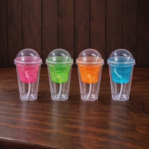 May include: Four clear plastic cups with colorful liquid and clear dome lids. The liquids are pink, green, orange, and blue. Each cup has a straw and is sitting on a wooden surface.