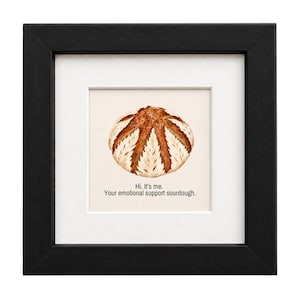 May include: A framed wall art piece featuring a centered illustration of a round, golden-brown sourdough loaf with a star-shaped score pattern. Below the bread, the text reads: Hi. It's me. Your emotional support sourdough. The artwork is enclosed in a black frame with a white mat.