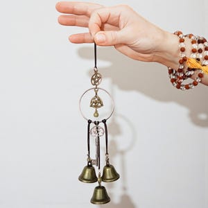 May include: A bronze-colored wind chime with a pentagram, Celtic knot, key, and three bells. The chime is suspended from a black cord. The background is a plain white wall.
