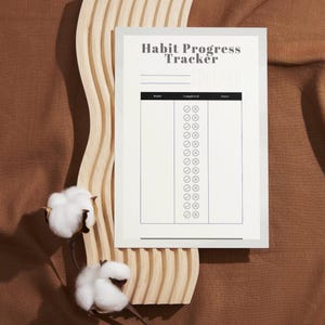 May include: A white habit progress tracker with the title at the top. The tracker has a section for habits, completed, and notes. The tracker is on a light wooden wavy surface, with cotton stems on the left side. The background is a brown fabric.