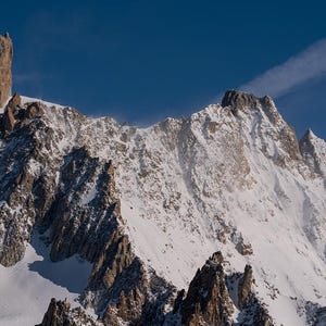 Może przedstawiać: Majestatyczny łańcuch górski pokryty śniegiem na tle czystego, błękitnego nieba. Obraz przedstawia poszarpane formacje skalne i charakterystyczną, wysoką iglicę. Scena oddaje poczucie rozległości i naturalnego piękna.