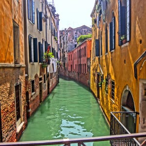 Könnte beinhalten: Eine malerische Kanalszene in Venedig, Italien, mit Gebäuden mit orangefarbenen, gelben und Backsteinfassaden. Die Gebäude haben dunkle Fensterläden und Fenster, und ein grüner Kanal verläuft zwischen ihnen. Ein Metallgeländer befindet sich im Vordergrund.