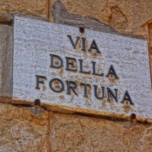 May include: A weathered street sign reading "VIA DELLA FORTUNA" is mounted on a textured, light brown stone wall. The sign is rectangular, with the Italian text in dark, bold letters. The stone has a rough, aged appearance.