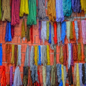May include: A vibrant display of colorful yarn bundles hanging against a red brick wall. The yarn is in various shades, including red, green, yellow, blue, and purple. The image showcases a variety of textures and hues, suggesting a craft or textile theme.