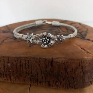 Könnte beinhalten: Silberfarbenes Armband mit floralem Design. Das Armband hat ein geflochtenes Band mit drei blütenförmigen Verzierungen. Die zentrale Blüte ist größer, mit einem strukturierten Zentrum, und die seitlichen Blüten sind kleiner. Das Armband wird auf einer Holzoberfläche präsentiert.