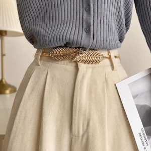 Peut inclure: Une ceinture en métal doré avec des accents en forme de feuilles. La ceinture est portée sur un pantalon crème et un cardigan gris. Le design de la ceinture présente deux grandes feuilles détaillées. L'esthétique générale est élégante et stylée.