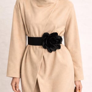 Op de afbeelding: Een beige wikkeljas met een zwarte elastische riem met een grote zwarte bloem. De jas heeft lange mouwen en een hoge kraag. De riem accentueert de taille en creëert een gedefinieerd silhouet. De jas is knielang.