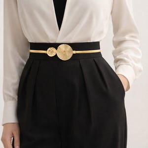 Peut inclure: Ceinture en métal doré avec deux éléments décoratifs circulaires. La ceinture est portée sur une ceinture noire, associée à un pantalon noir et un chemisier blanc. Le design de la ceinture ajoute une touche d'élégance à la tenue.