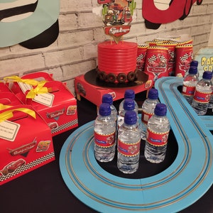 May include: A Cars-themed birthday party table setting with a red cake, blue race track, and water bottles with Cars character labels. The table is decorated with red and yellow boxes, and a stop sign.
