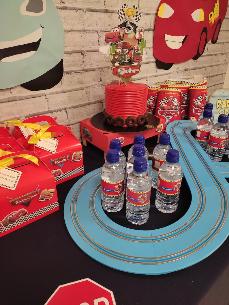May include: A Cars-themed birthday party table setting with a red cake, blue race track, and water bottles with Cars character labels. The table is decorated with red and yellow boxes, and a stop sign.