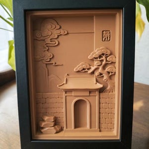 May include: A framed relief sculpture depicting an Asian-inspired landscape. The artwork features a temple, clouds, and a tree, all in a warm brown hue, set against a textured background. The frame is black.