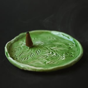 Op de afbeelding: Een groene keramische wierookhouder met een bladeren ontwerp. Een bruine wierookkegel staat in het midden en geeft een wolkje rook af. De houder heeft een licht onregelmatige rand en staat tegen een donkere achtergrond.