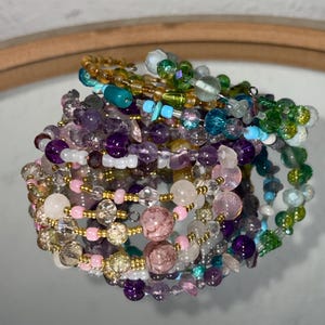 May include: A collection of beaded bracelets in various colors, including purple, pink, green, and gold. The bracelets are made with a variety of beads, including gemstones and glass beads. The bracelets are stacked together, creating a layered look.